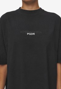 Svart bomullströja med rund hals, med broderad text "PECADOR" och "PGDR" i vitt, över bröstet. Korta ärmar, avslappnad passform.