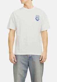 T-shirt bianca in cotone con maniche corte e collo rotondo. Presenta un design grafico blu sulla parte sinistra del petto. Vestibilità comoda, texture morbida.