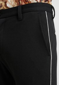 Schwarze Hose aus strukturiertem Stoff mit einem weißen Paspelierungsdetail an der Seite. Mit einer Vordertasche und einem flachen Bund.