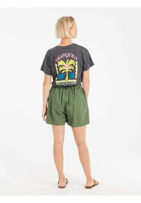 Grijs t-shirt met een kleurrijke palmboomprint; groene shorts met een elastische tailleband; platte sandalen; van achteren gezien tegen een witte achtergrond.