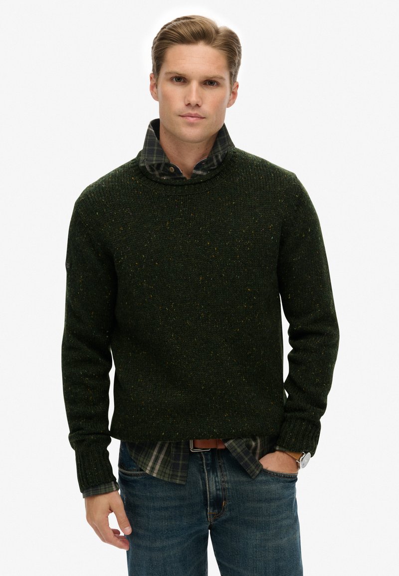 Pull en tricot vert avec des taches, design à col rond, associé à une chemise à carreaux avec col en dessous. Porté avec un jean bleu et une montre.