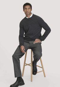 Dunkelgrauer Strickpullover mit rundem Hals, kombiniert mit maßgeschneiderten grauen Hosen und schwarzen Knöchelstiefeln, sitzt auf einem Holzschemel.