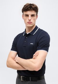 Νεαρός άνδρας με σταυρωμένα χέρια, φορόντας σκούρο μπλε πόλο πουκάμισο με λευκή διακόσμηση και το λογότυπο "BOSS" στο στήθος.