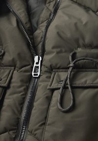 Veste rembourrée vert olive avec une fermeture éclair argentée, cordon de serrage ajustable et deux poches avant avec fermetures à pression.