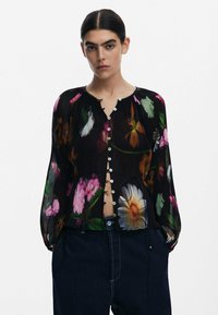 Transparent svart blus med färgglad blommönster, knäppning fram och elastiska ärmslut; har plisserad textur och dragsnöre i nederkant.
