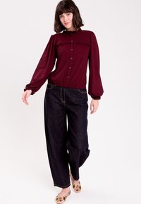 Burgunderfarbene Bluse mit langen Ärmeln, Volantkragen und -manschetten, kombiniert mit weit geschnittenen, dunkelblauen Jeans und blumenmusternden Schuhen.