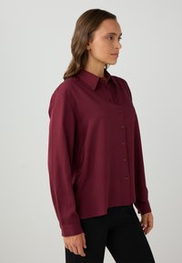 Femme portant une blouse bordeaux à manches longues avec des boutons, associée à un pantalon noir, se tenant de profil contre un fond clair.