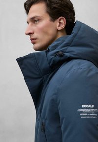 Granatowa ocieplana kurtka z wysokim kołnierzem, regulowanym kapturem, kieszeniami na zamek oraz brandingiem Ecoalf na rękawie. Gładka, matowa struktura.