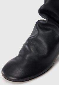 Botte en cuir noire avec une texture lisse et un bout arrondi. Présente un tissu doux et froissé sur le dessus pour un aspect décontracté.