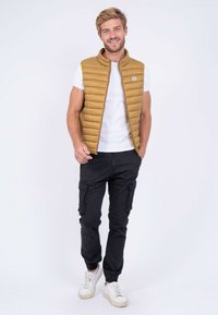 Gilet matelassé beige avec un quilting horizontal, T-shirt blanc, pantalon cargo noir et baskets blanches. Le gilet présente un logo rond sur la poitrine.