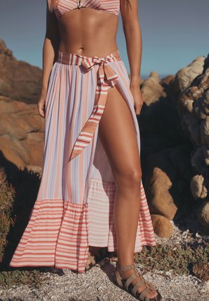Kvinde iført en stribet bikini top og matchende nederdel med høj slids og sandaler, står på klippefyldt terræn under klar himmel.