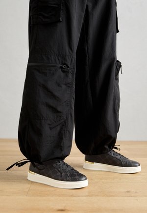 Pantalones cargo negros con bolsillos con cremallera y cordones en los puños, combinados con zapatillas negras de caña baja con suelas blancas y detalles dorados.