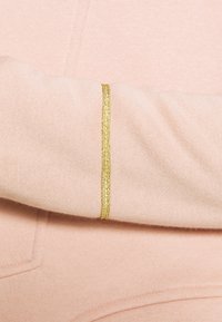 Matière de sweat-shirt rose clair avec une texture lisse et une bande métallique dorée le long du poignet. Design simple, sans motifs.