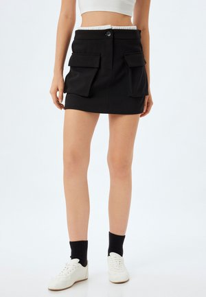 Zwarte minirok met een aansluitend ontwerp, voorzien van twee grote zakken aan de voorkant, een knoopsluiting en een elastische taille. Gepaard met witte sneakers.