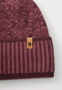 Knit beanie in shades of burgundy and deep maroon, featuring a ribbed brim and a leather tag with logo. Soft texture and patterned top.