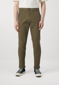 Olivenfargede bukser med rett passform, knappelukking og oppbrettede ermer, kombinert med svarte sneakers med hvit såle og snøring.
