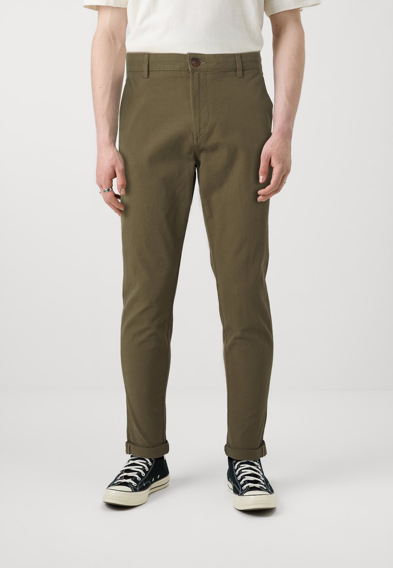 Olivenfargede bukser med rett passform, knappelukking og oppbrettede ermer, kombinert med svarte sneakers med hvit såle og snøring.