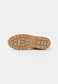 Suola di un boot marrone, robusto, con profondi modelli di battistrada e perforazioni circolari su uno sfondo bianco semplice.