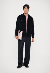 Crna kordna overshirt, svijetloružičasta majica, tamnoplave hlače s prugicama, drži malu ružičastu torbicu, u pozadini jednostavna bijela pozadina.