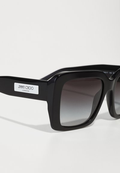 Schwarze übergroße Sonnenbrille mit glänzender Oberfläche, ausgestattet mit einem Farbverlauf-Glas und einem weißen Logo-Akzent am Bügel. Rechteckiges Rahmendesign.