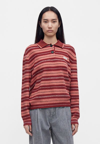 Jeune femme aux longs cheveux noirs portant un pull à manches longues rayé rouge et rose avec deux boutons et un pantalon gris sur un fond uni.