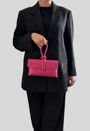 Borsa a mano in pelle rosa con manico annodato e chiusura a patta. La superficie è liscia ed è tenuta da una persona con blazer nero.