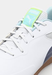 White sports shoe with a textured upper, pastel blue accents, a green tag with "PUMA," and a brown rubber sole. Laced design.