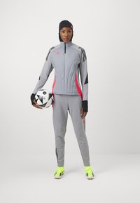 Šedá sportovní bunda s růžovými akcenty a logem, doplněná o odpovídající kalhoty. Hráč drží černobílý fotbal, obutý má jasně žluté kopačky.