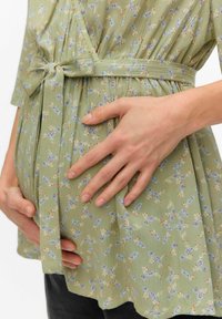 Zeleni cvetni top s kratkimi rokavi, ki ima oblečen pas, teksturirano tkanino in majhne modre ter rdeče cvetlične vzorce po površini.