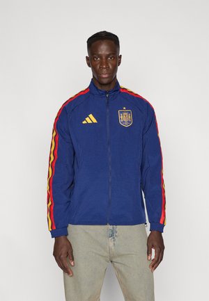 Veste de sport bleue avec fermeture éclair, présentant des accents rayés rouges et jaunes sur les manches, un logo brodé Espagne et la marque Adidas sur la poitrine.
