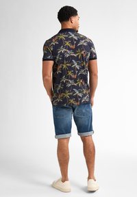 Camisa polo azul marino con un patrón tropical de palmeras, combinada con pantalones cortos de denim enrollados. Un calzado casual completa el look.