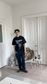 Svart t-skjorte med grafisk design, blå jeans og mørke sko. Står i et rom med hvite vegger og tregulv.