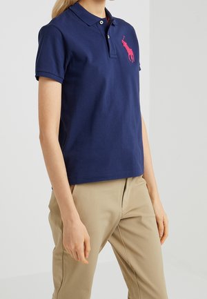 Kvinde iført en mørkeblå polo med en lyserød broderet logo og beige bukser mod en ensfarvet lys baggrund.