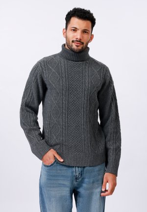 Suéter de cuello alto de punto trenzado gris oscuro con patrones texturizados, puños y bajo acanalados. Lleva puesto jeans azul claro, con las manos en los bolsillos.