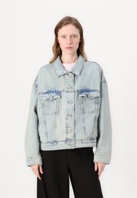 Light blue denim jacket with a relaxed fit, featuring double breast pockets, button closure, and subtle embossed lettering on the fabric.