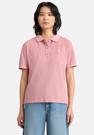 Mujer de cabello negro con una camisa polo rosa de manga corta y jeans azul claro, posando frente a un fondo sencillo.