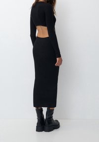 Robe noire à manches longues avec un dos découpé, silhouette ajustée, texture douce et coupe mi-longue, associée à des bottes noires épaisses.