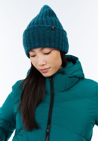 Moteris, užsidėjusi žalia megztą kepurę ir atitinkantį žalią pūkinę striukę su kapišonu, žiūri žemyn priešais paprastą baltą foną.