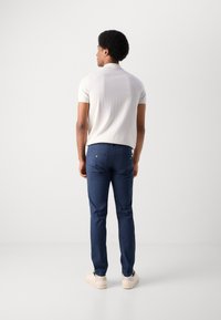 Polo shirt blanc à manches courtes et pantalon ajusté bleu foncé, affichant une coupe slim, une texture lisse et un minimalisme, porté avec des chaussures claires.