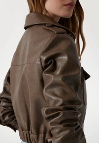 Giacca in pelle marrone con una vestibilità rilassat, grande colletto e maniche gather. Presenta una texture liscia e una leggera lucentezza, con una tasca laterale.