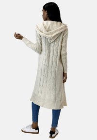 Stickad kofta i elfenben med huva, intrikat kabelstickad mönster, ribbstickade muddar och fåll. Matchas med blåa smala jeans och sneakers.