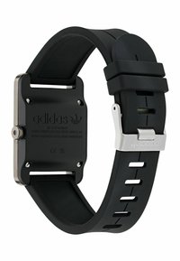 Montre de sport en caoutchouc noir avec un cadran carré, des éléments en métal argenté, et un bracelet texturé comportant des trous pour un ajustement réglable. Logo embossé sur le bracelet.