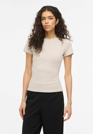 Žena s dlhými kučeravými vlasmi, oblečená v priliehavom béžovom tričku s krátkym rukávom a čiernych nohaviciach, stojaca pred jednoduchým bielym pozadím.