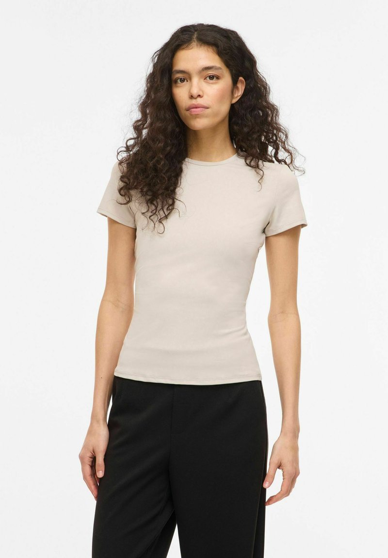 Kvinne med langt, krøllete hår, iført en tettsittende beige topp med korte ermer og svarte bukser, stående mot en enkel hvit bakgrunn.