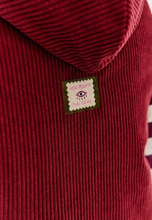 Czerwona bluza z korduroyu z wyszywaną łatą przedstawiającą oko i napisem "Zabłyśnij jaśniej niż słońce."
