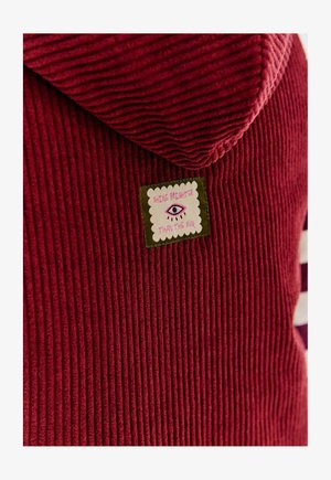 Czerwona bluza z korduroyu z wyszywaną łatą przedstawiającą oko i napisem "Zabłyśnij jaśniej niż słońce."