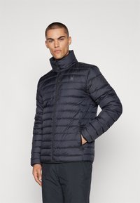 Mann in einer schwarzen, gesteppte Jacke mit hohem Kragen, der mit einer Hand in der Tasche vor einem schlichten Hintergrund steht.