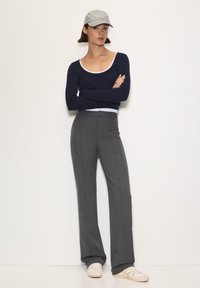 Marineblauw top met lange mouwen over een wit shirt, gecombineerd met grijze broek met wijde pijpen en crèmekleurige sneakers, aangevuld met een lichtgrijze pet.