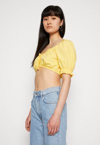 Top amarelo cropped com mangas bufantes, punhos elásticos e detalhe de laço na frente; combinado com jeans claros de cintura alta. Textura de algodão suave.