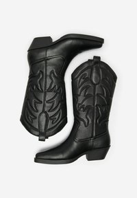 Botas de vaquero de cuero negro con costuras decorativas, punta afilada y un tacón bajo en forma de bloque. Presentan un patrón en forma de llama en el cañón.